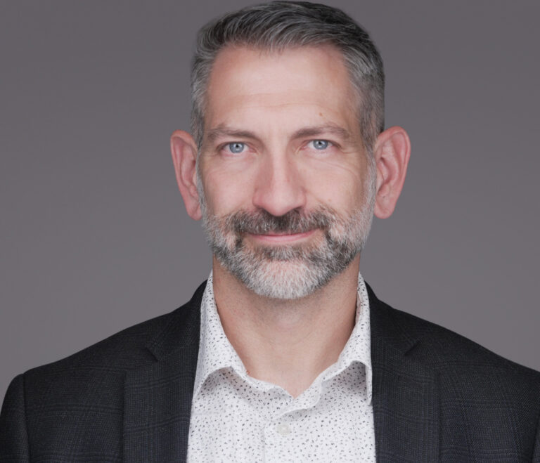 Professional headshot of a smiling man with a gray beard wearing a black blazer and patterned white shirt