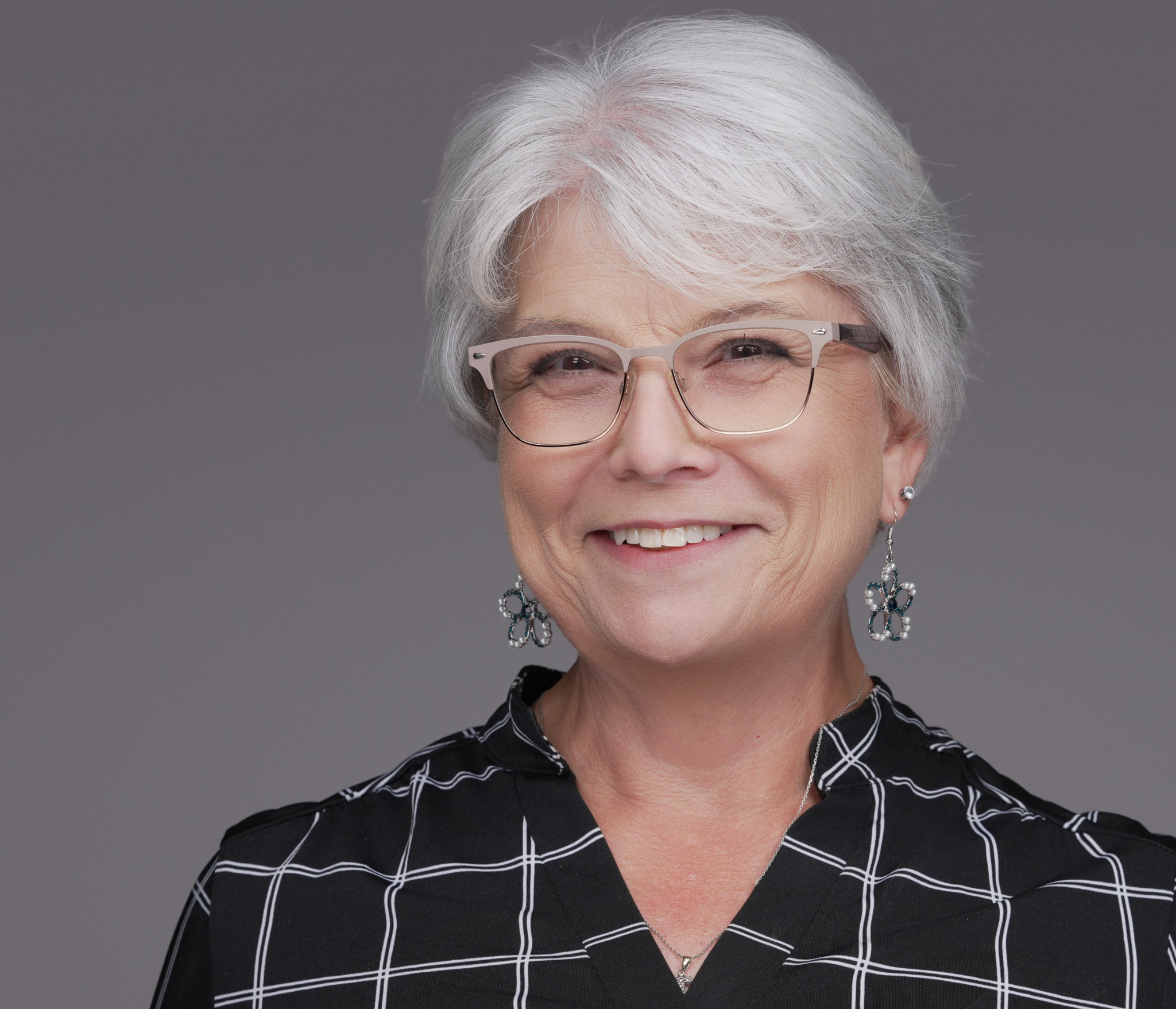 Smiling woman with short gray hair and glasses in a plaid top, studio headshot against a gray backdrop
