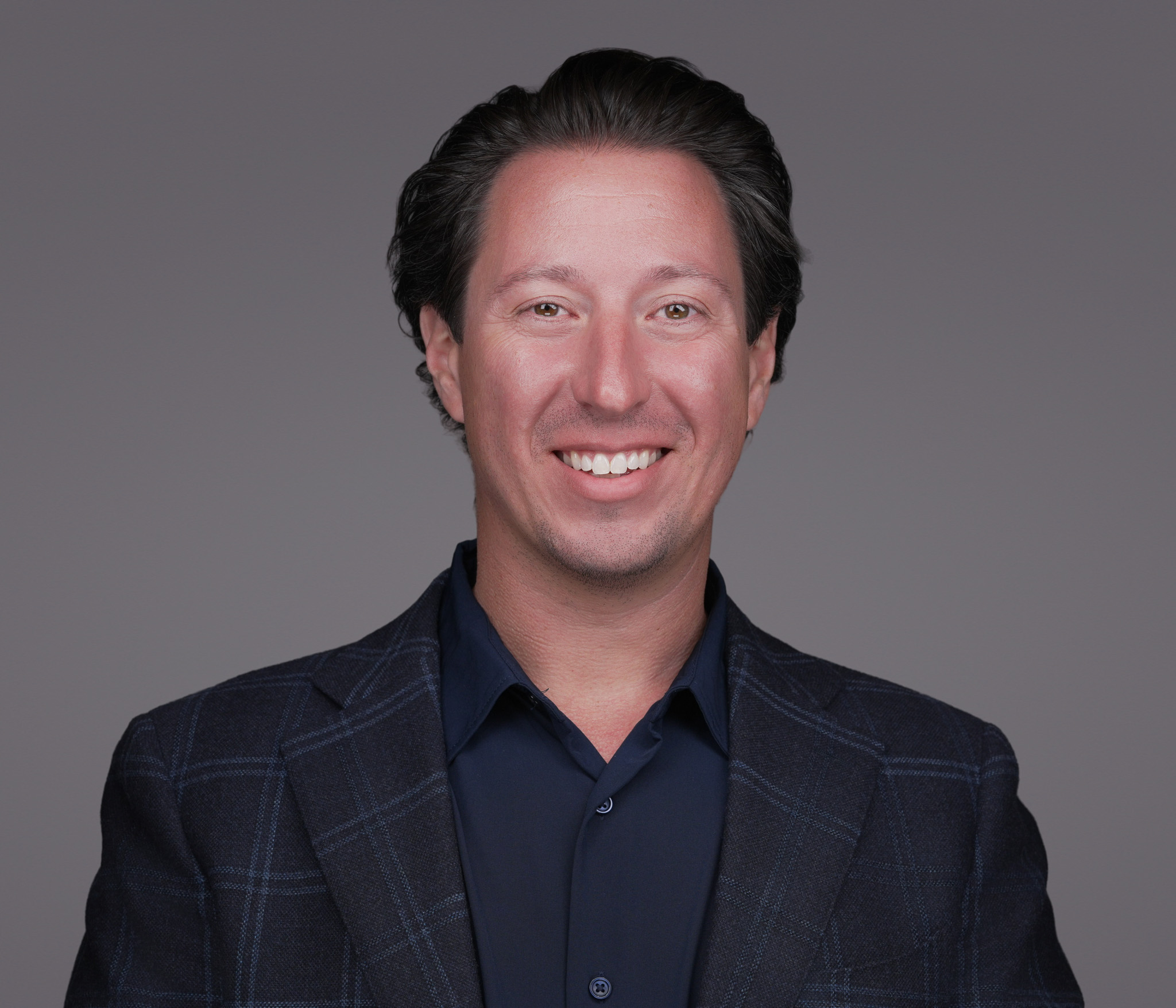 Smiling man with dark hair in a navy blazer, studio headshot against a dark gray backdrop