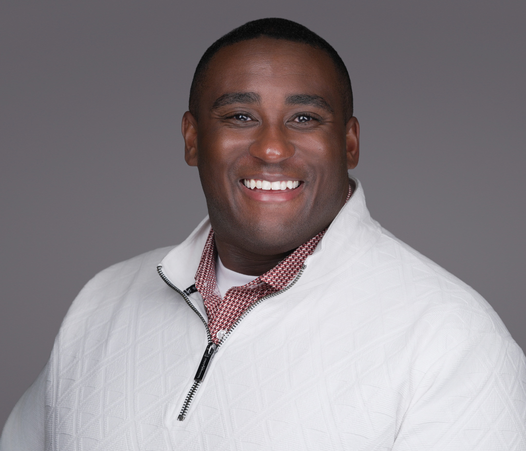 Smiling man in a white zip-neck sweater, studio headshot against a dark gray backdrop