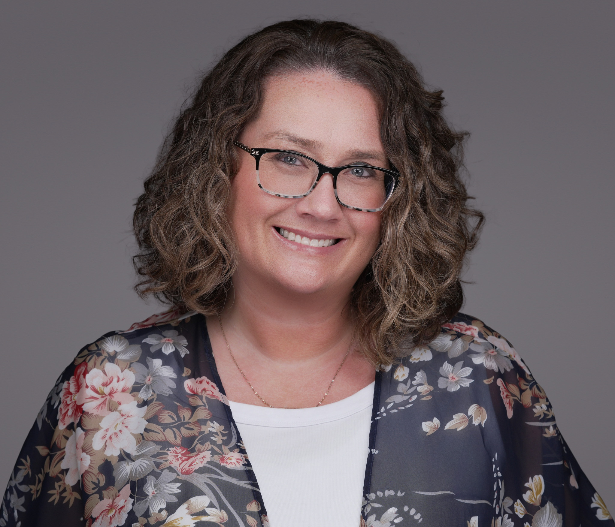Smiling woman with curly hair and glasses in a patterned blazer, studio headshot against a dark gray backdrop
