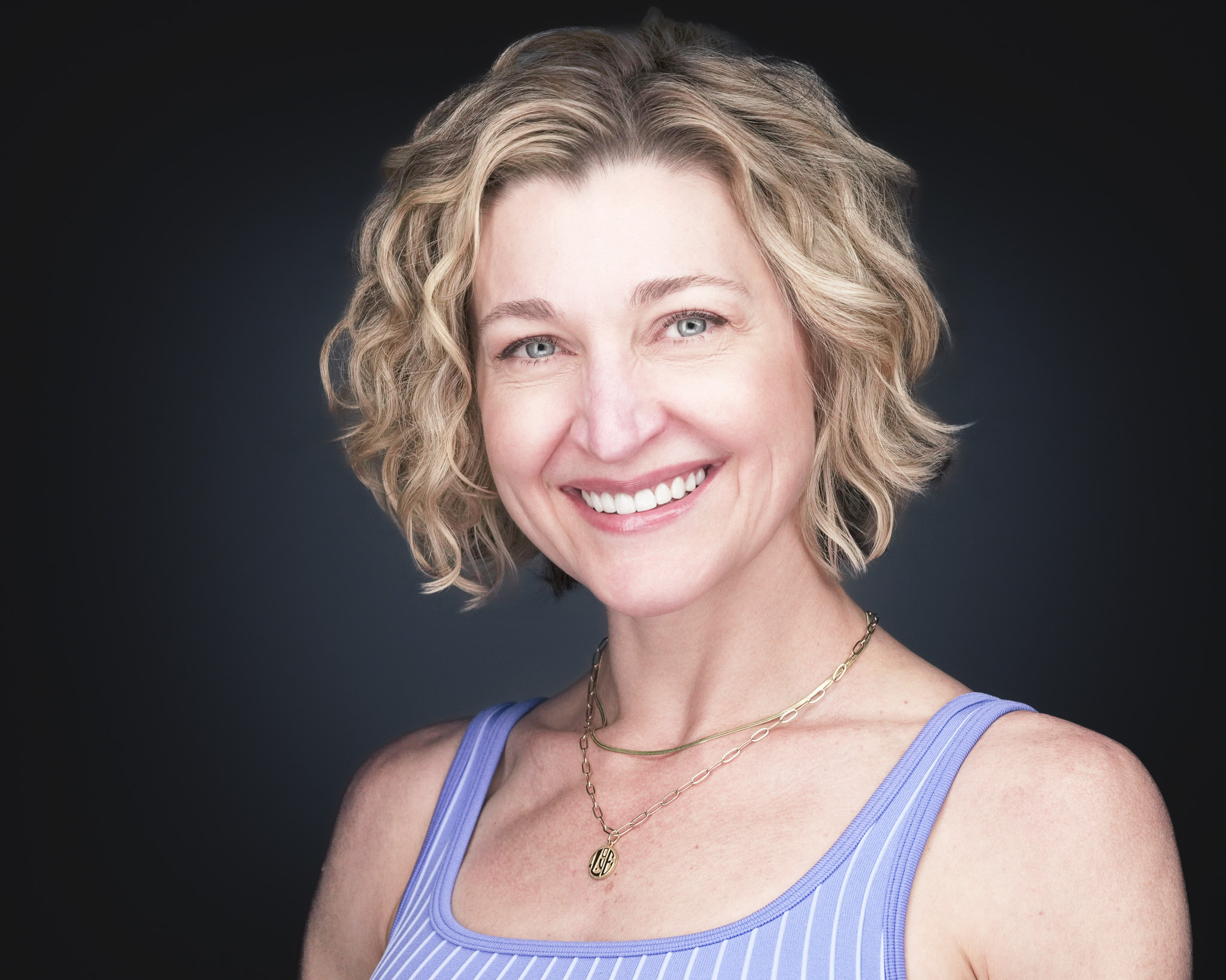 Smiling woman with wavy blonde hair in a lavender top, studio headshot against a dark gray backdrop