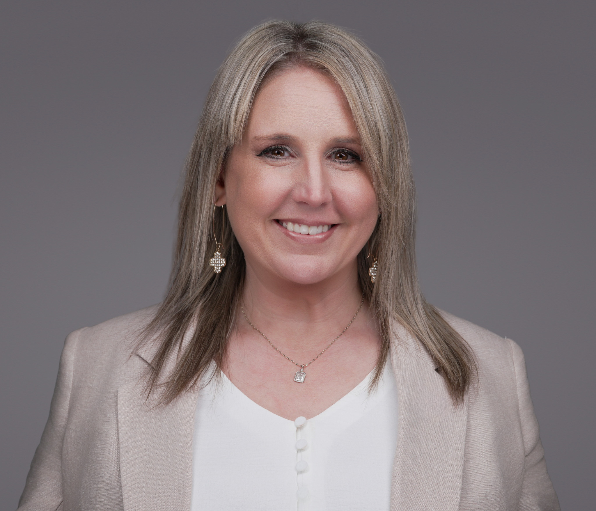 Smiling woman with long blonde hair in a beige blazer, studio headshot against a dark gray backdrop