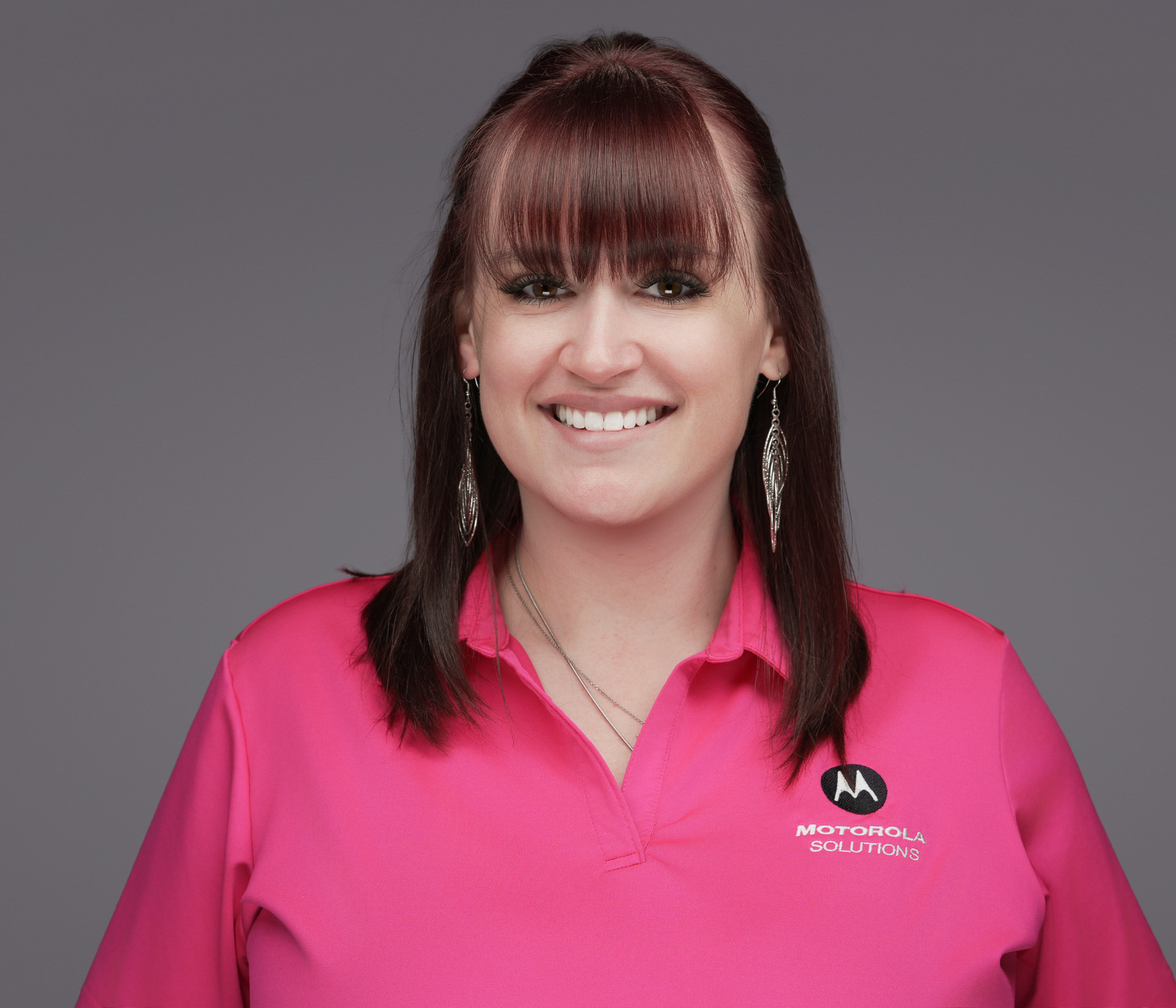 Smiling woman with dark hair and bangs in a pink top, studio headshot against a dark gray backdrop