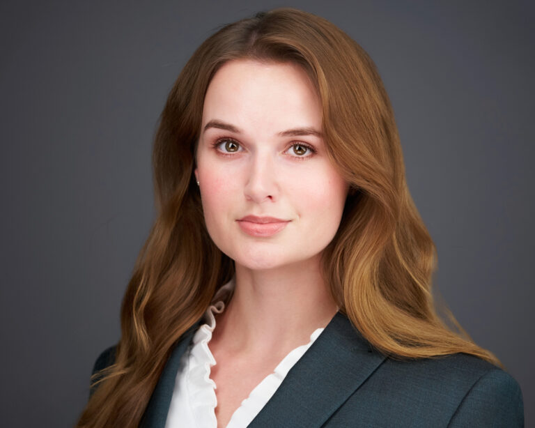 Professional headshot of a woman with long wavy auburn hair wearing a dark green blazer against a gray studio background
