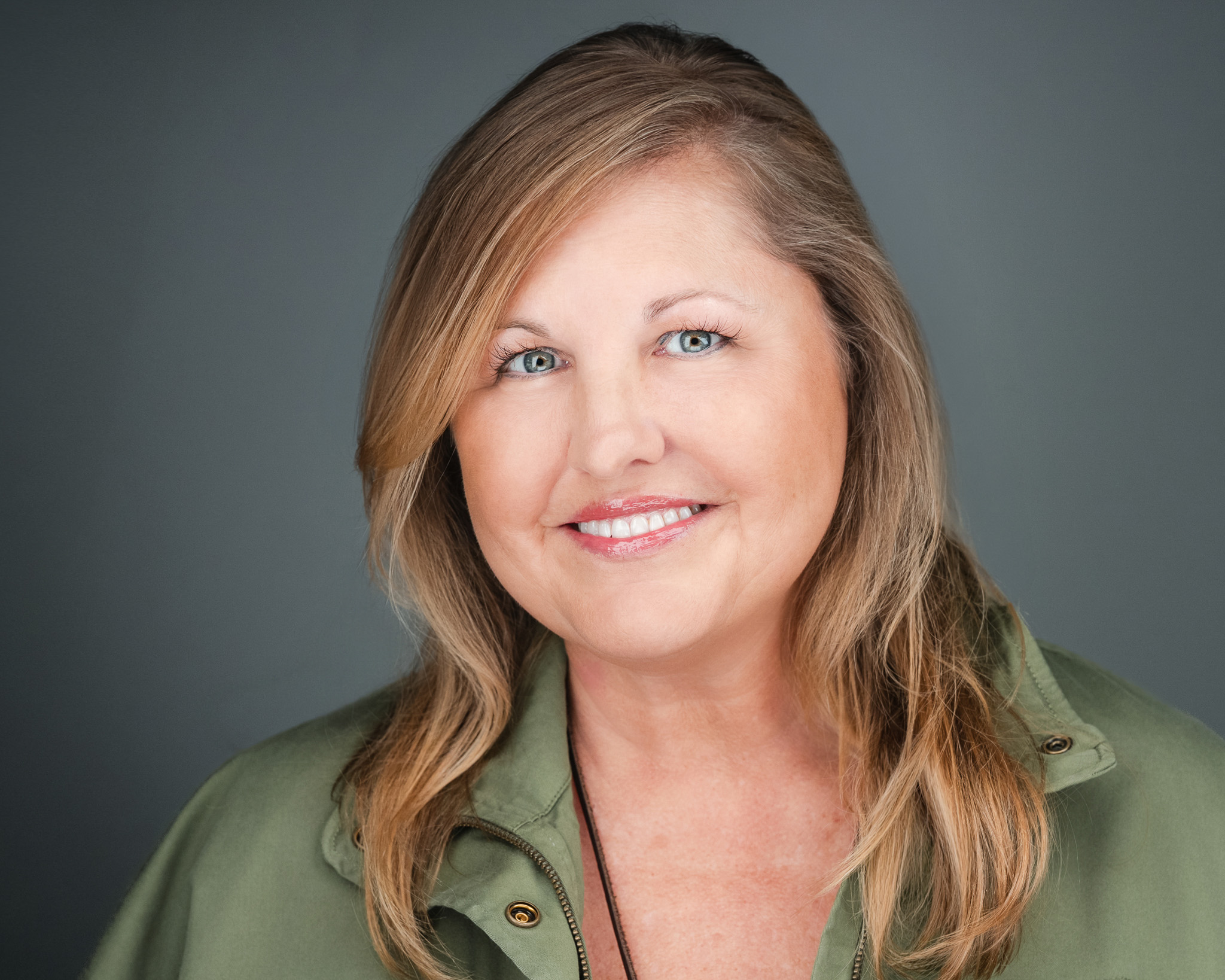 Smiling woman with long blonde hair in an olive green top, studio headshot against a blue-gray backdrop