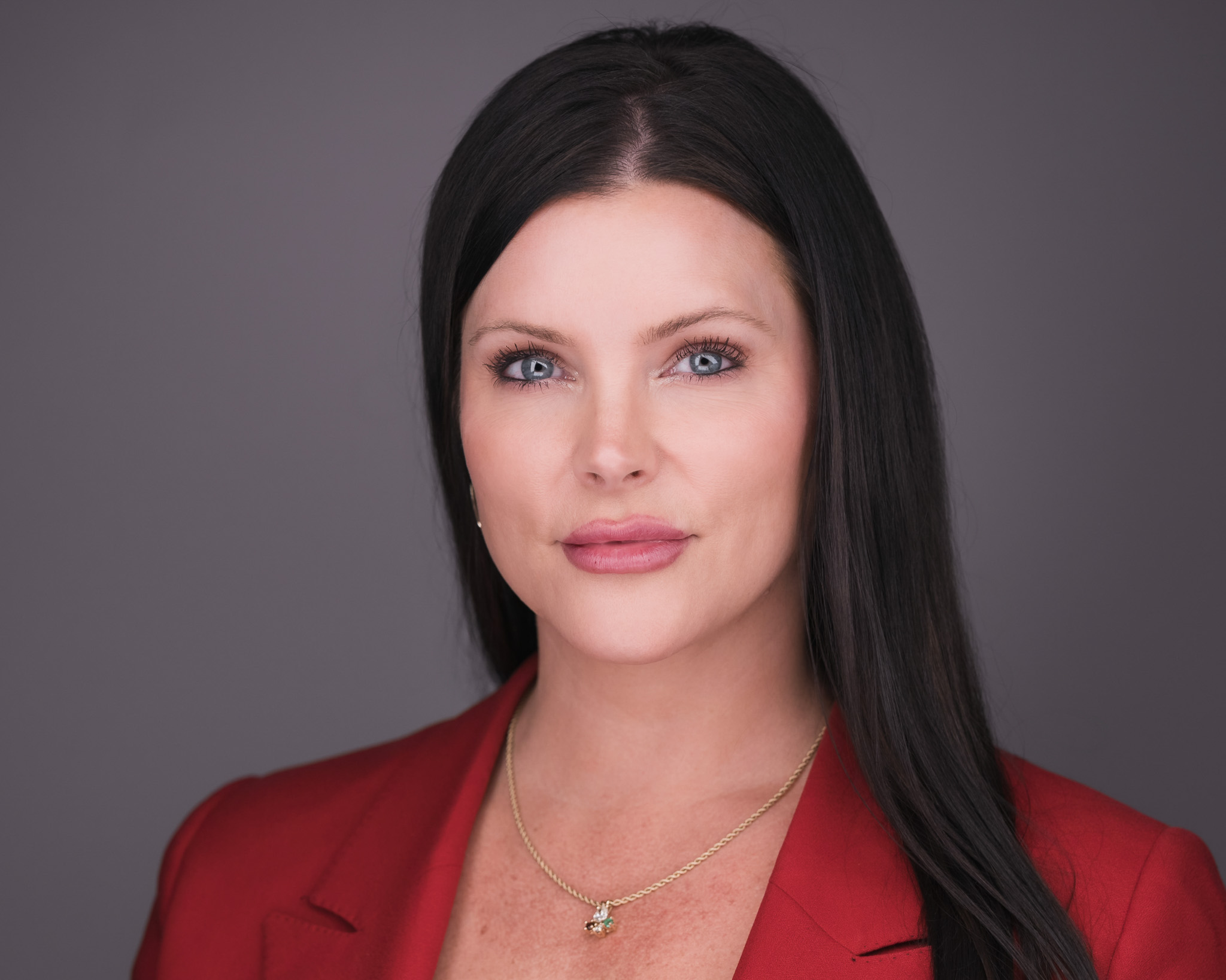 Woman with long black hair in a red blazer, studio headshot against a gray backdrop