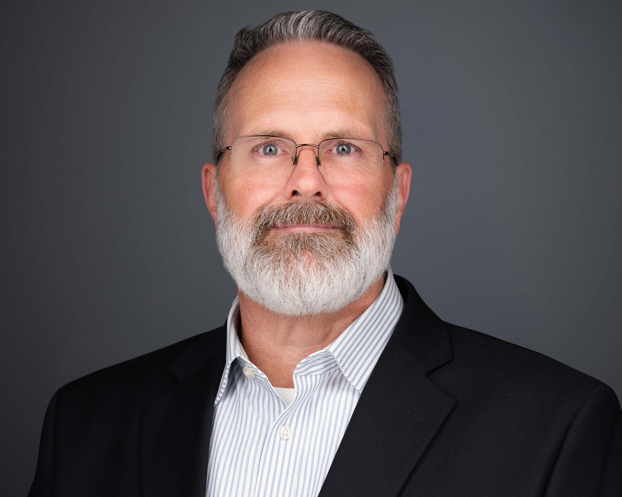 Man with a gray beard and glasses in a dark blazer, studio headshot against a dark gray backdrop