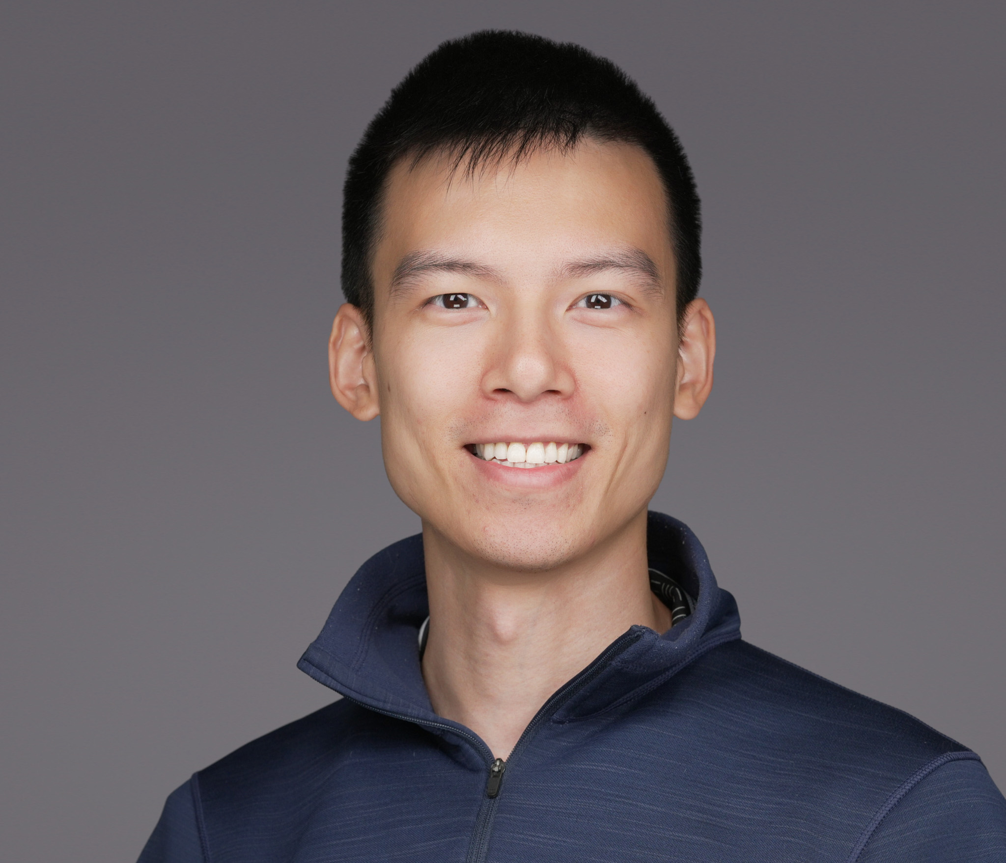 Smiling man with dark hair in a navy sweater, studio headshot against a dark gray backdrop