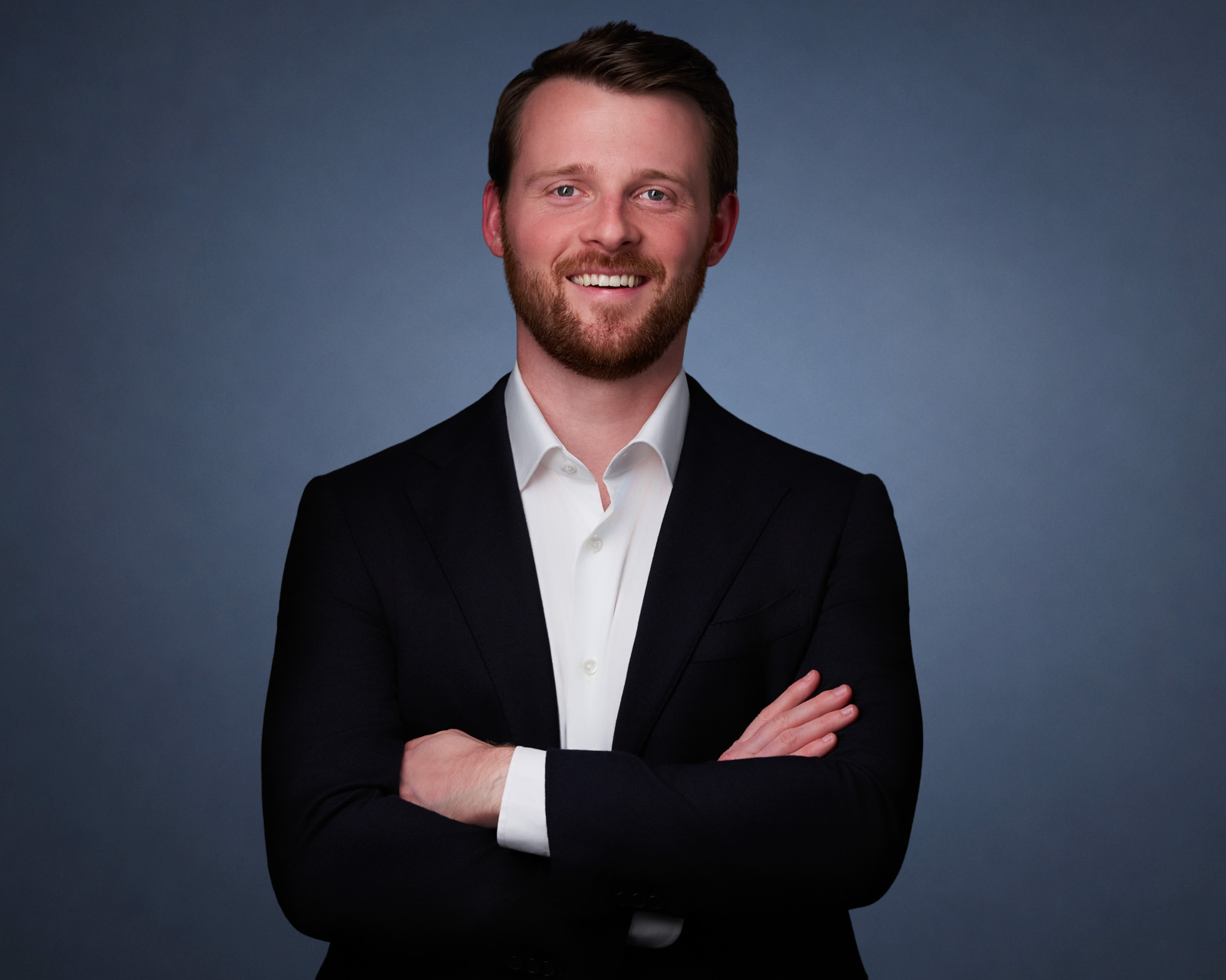 Man in a dark blazer with arms crossed, studio headshot against a blue-gray backdrop