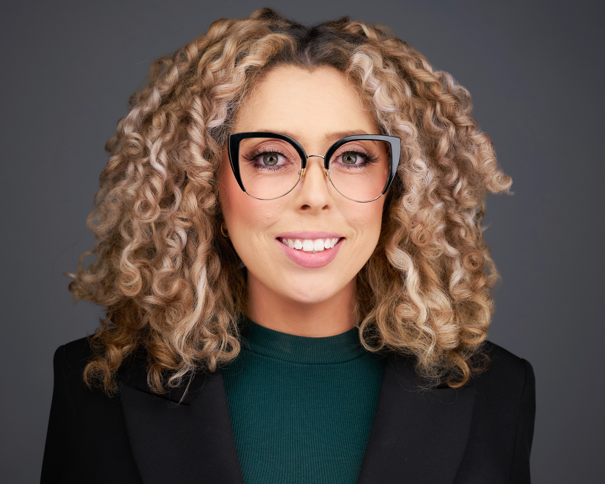 Smiling woman with curly hair and glasses in a dark blazer, studio headshot against a dark gray backdrop