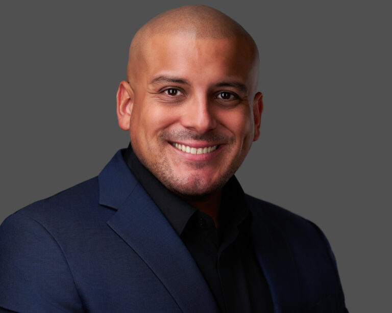 Professional headshot of a smiling bald man wearing a navy blazer over a black shirt against a gray background